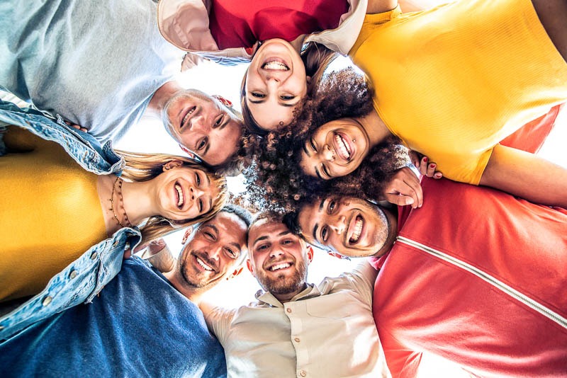 Multiracial group of young people standing in circle and smiling at camera - Happy diverse friends having fun hugging together - Low angle view